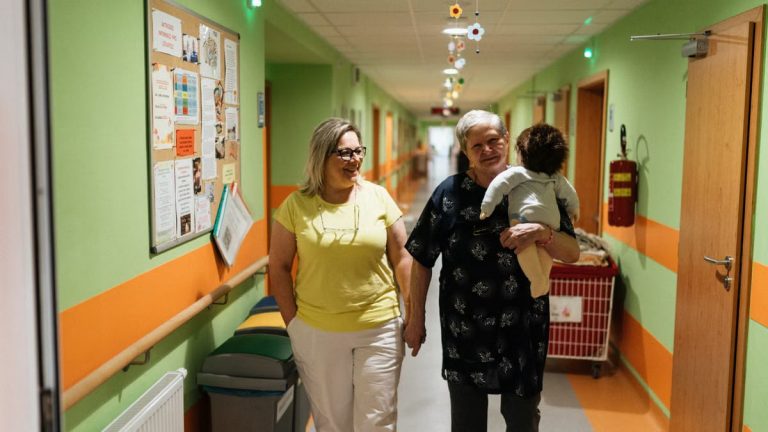 Two women walking in a nursing home corridor, one holding a child doll.