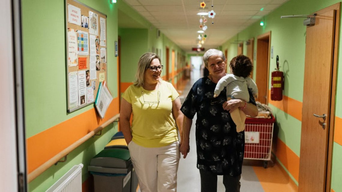 Two women walking in a nursing home corridor, one holding a child doll.