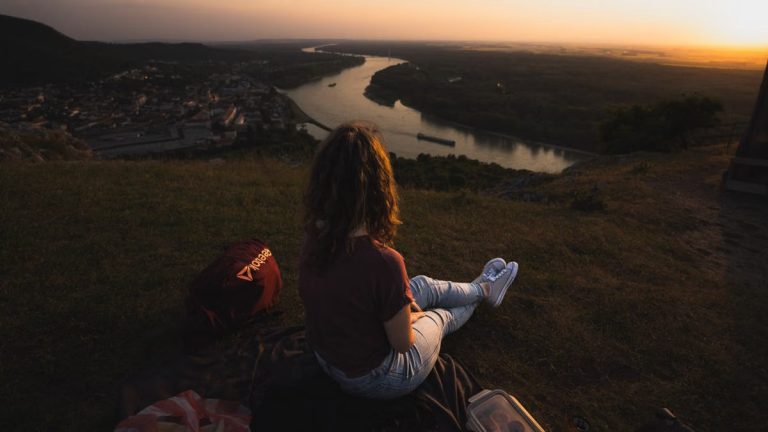 Woman sits on hillside overlooking river and town at sunset, creating a serene landscape scene.